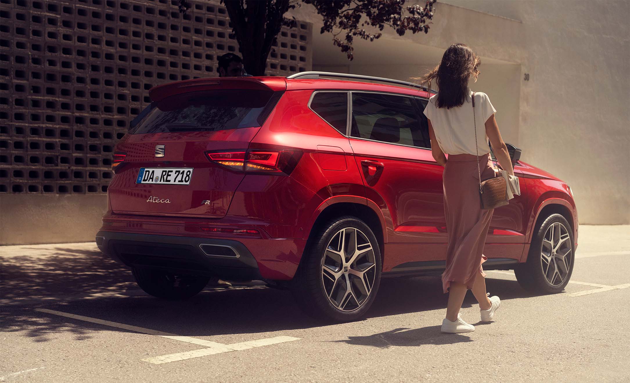 Eine Frau geht auf einen am Straßenrand geparkten roten SEAT Ateca FR zu, im Hintergrund eine helle Wand mit Strukturmuster und ein Baum im Schatten.