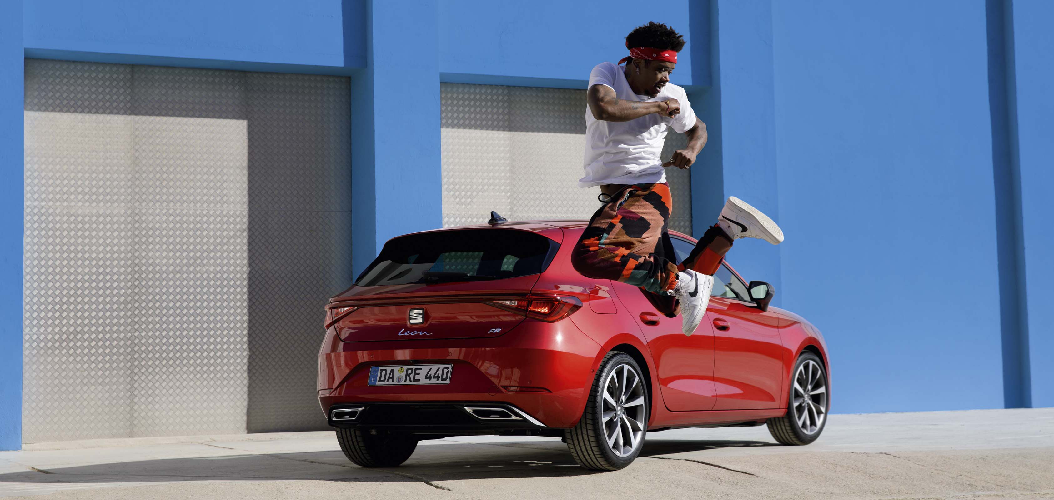 Ein roter SEAT Leon steht vor einer blauen Wand, Seitenansicht. Vor ihm tanzt ein Mann mit roter Hose und weißem T-Shirt.