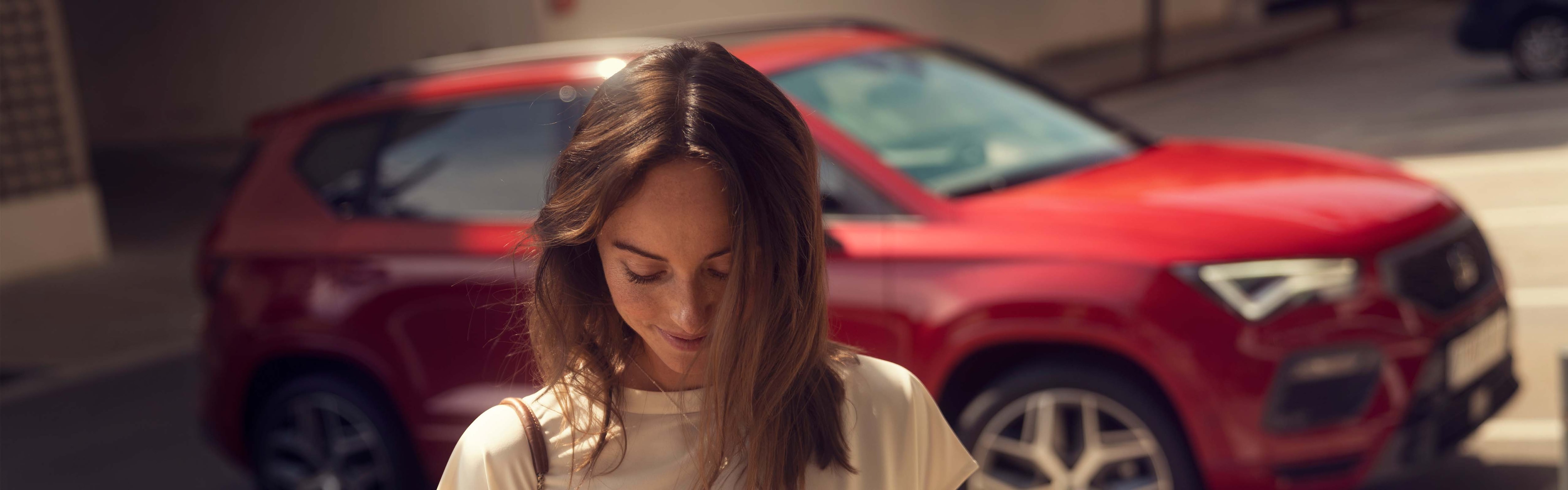 Eine Frau blickt auf ihr Smartphone, während im Hintergrund ein roter SEAT Ateca FR in einer Parklücke steht.