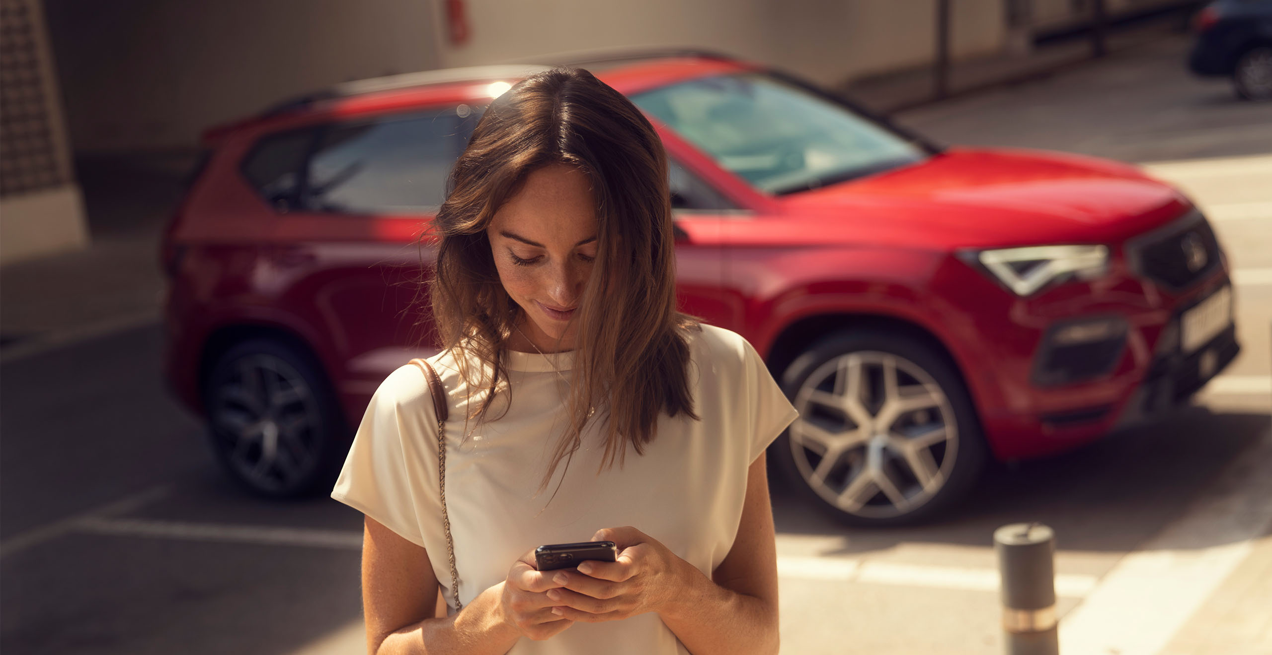 Eine Frau blickt auf ihr Smartphone, während im Hintergrund ein roter SEAT Ateca FR in einer Parklücke steht.