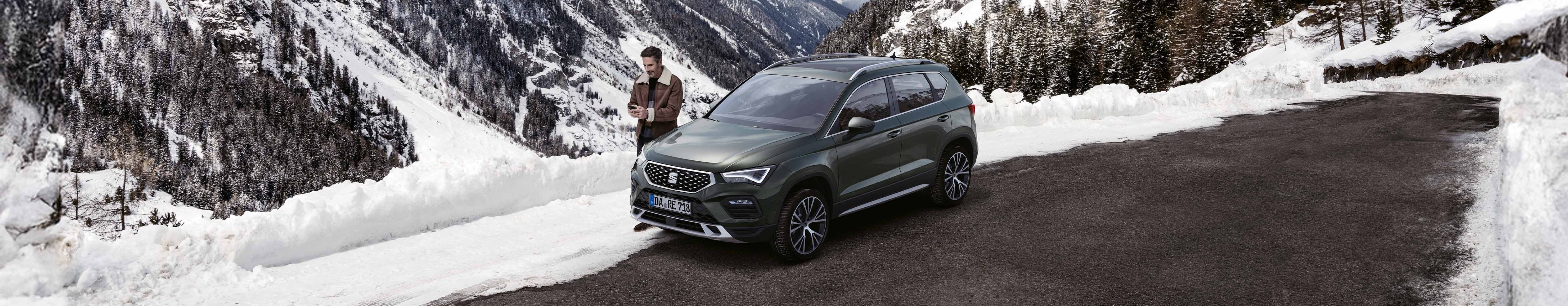 Ein SEAT Ateca SUV parkt auf verschneitem Boden, daneben stehen zwei Personen mit Snowboards. Winterkompletträder 2024 von SEAT, ideal für verschneite Straßen.