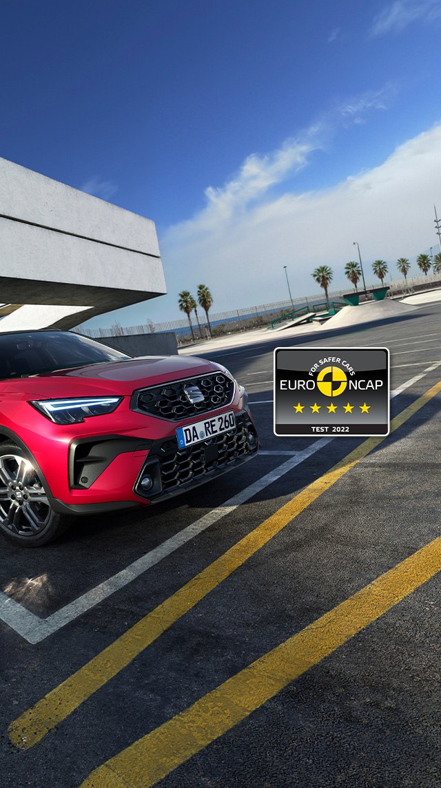 Roter SEAT Arona von hinten auf einem Parkplatz, modernes Gebäude im Hintergrund. Nummernschild mit deutscher Kennung, fünf Sterne Euro NCAP Logo unten rechts. Parkstreifen und rote Geländer im Vordergrund