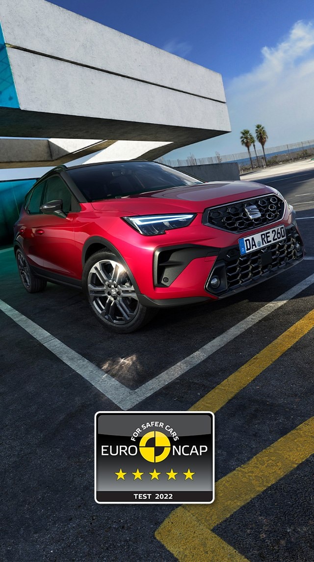 Roter SEAT Arona von hinten auf einem Parkplatz, modernes Gebäude im Hintergrund. Nummernschild mit deutscher Kennung, fünf Sterne Euro NCAP Logo unten rechts. Parkstreifen und rote Geländer im Vordergrund