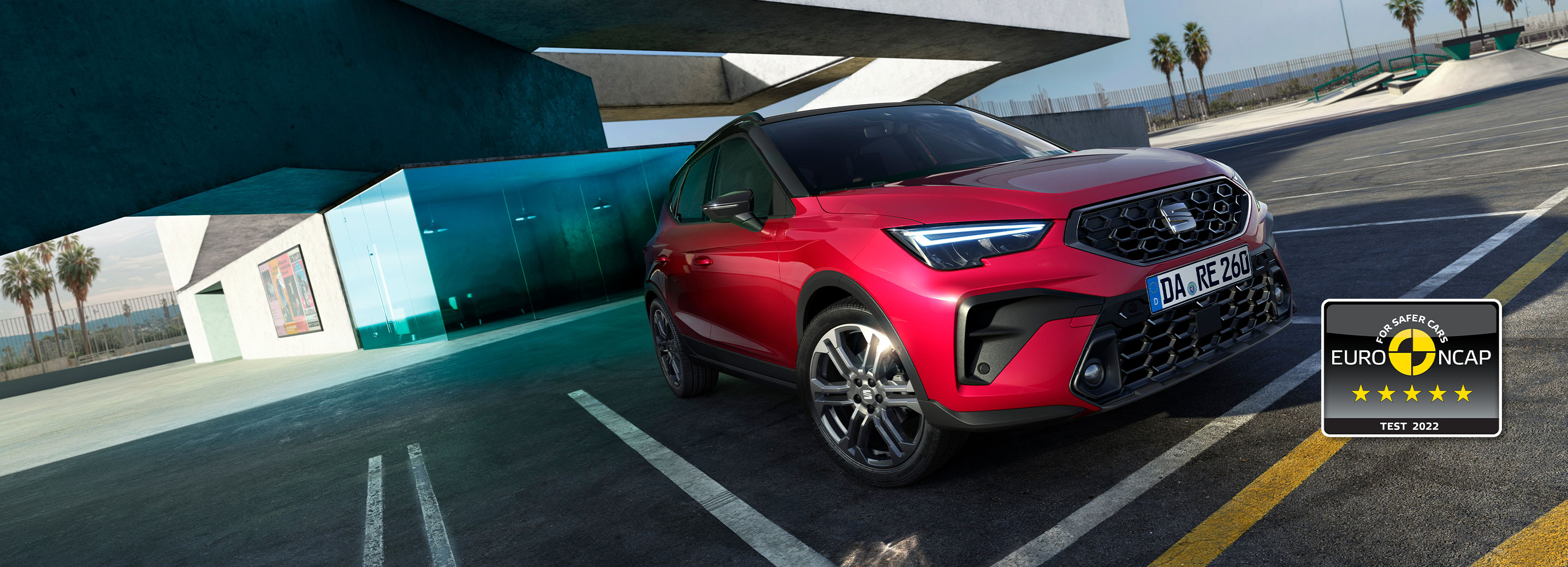 SEAT Arona SUV in Frontansicht auf einem Parkplatz, gestaltete Front mit hexagonalem Kühlergrill, LED-Scheinwerfer, zweifarbiges Design vor einem Gebäude mit großer Glasfassade, Palmen im Hintergrund (Variante)