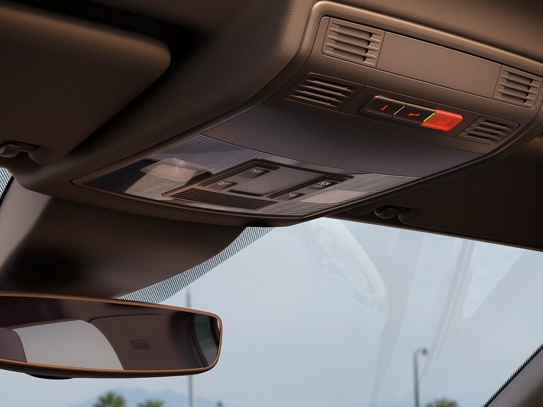 Innenansicht eines Autos mit Fokus auf den Rückspiegel und die Beleuchtungseinheit an der Decke. Im Hintergrund ist durch die Windschutzscheibe eine moderne Architektur mit Beton und roten Elementen zu sehen