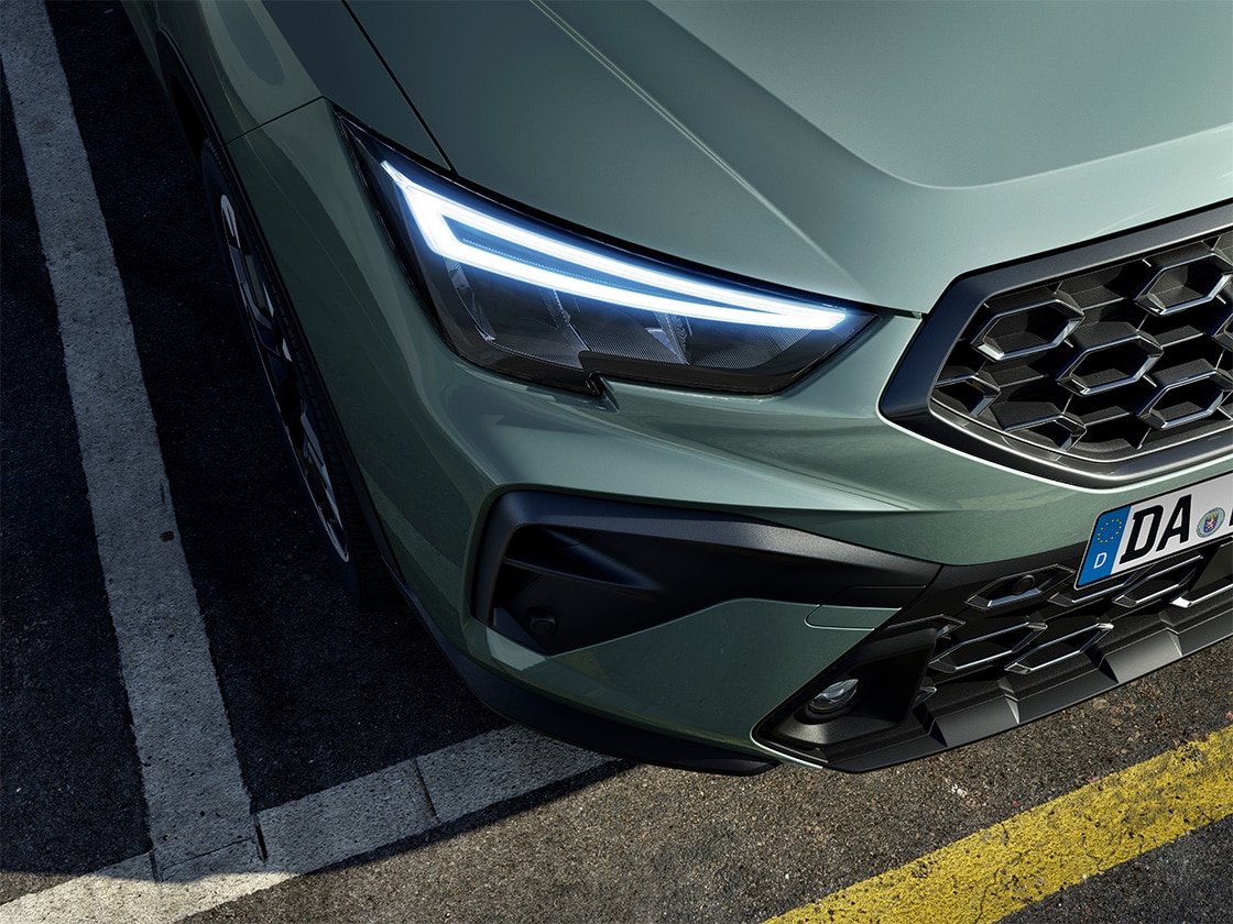 Frontansicht eines grünen SEAT Arona mit modernen LED-Scheinwerfern und markantem Kühlergrill. Detailaufnahme auf einem Parkplatz mit gelben und weißen Markierungen am Boden. Nummernschild mit "DA" sichtbar.