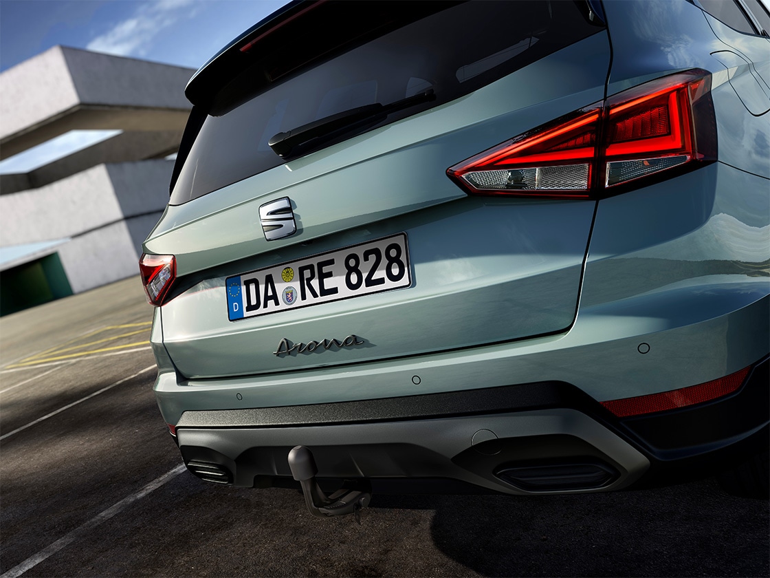 Rückansicht eines grünen SEAT Arona mit erkennbaren Rückleuchten und Anhängerkupplung vor einem modernen Gebäude. Fahrzeugemblem und Modellname sind am Heck sichtbar. Nummernschild mit der Aufschrift "DA RE 828".