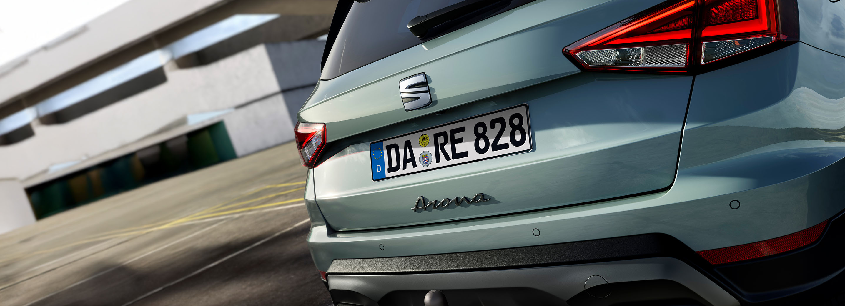 Rückansicht eines grünen SEAT Arona mit erkennbaren Rückleuchten und Anhängerkupplung vor einem modernen Gebäude. Fahrzeugemblem und Modellname sind am Heck sichtbar. Nummernschild mit der Aufschrift "DA RE 828".