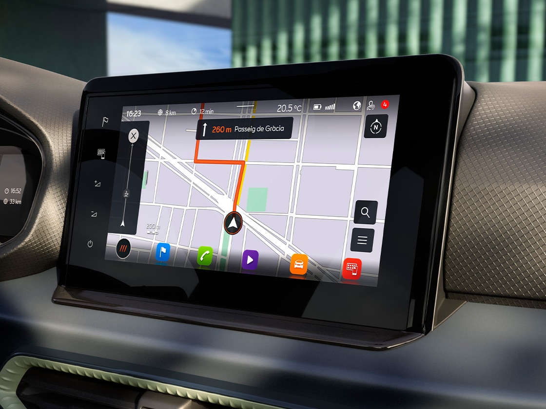 Navigationsbildschirm in einem Auto zeigt eine Routenanleitung zum Passeig de Gracia mit orangefarbener Linie. Weitere Apps und Funktionen am unteren Rand der Anzeige sichtbar. Der Bildschirm ist in die Armatur integriert.