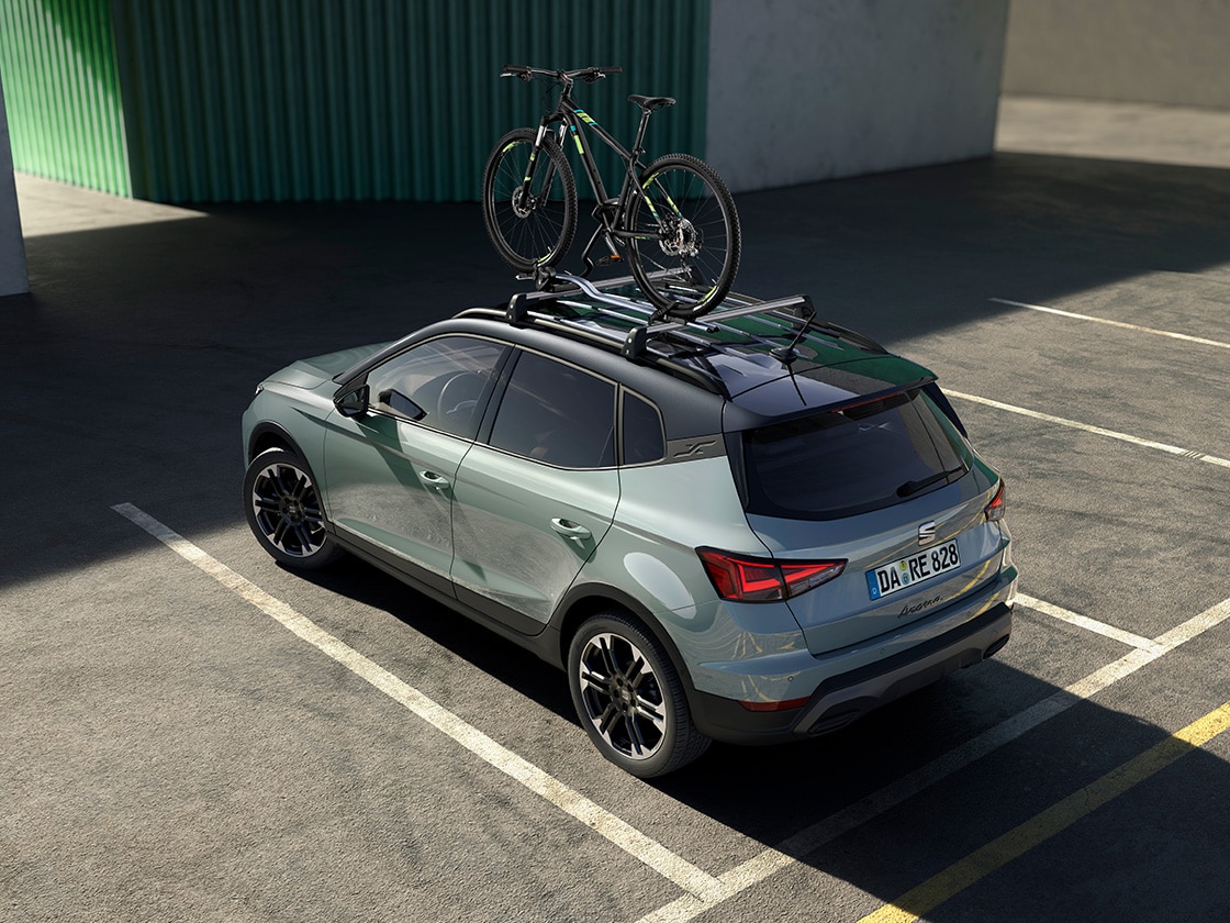 SEAT Arona in grau von schräg oben in einem leeren Parkplatz. Fahrrad auf dem Dachträger montiert, grüne Gebäudewand im Hintergrund. Nummernschild mit der Aufschrift "DA RE 828" sichtbar.