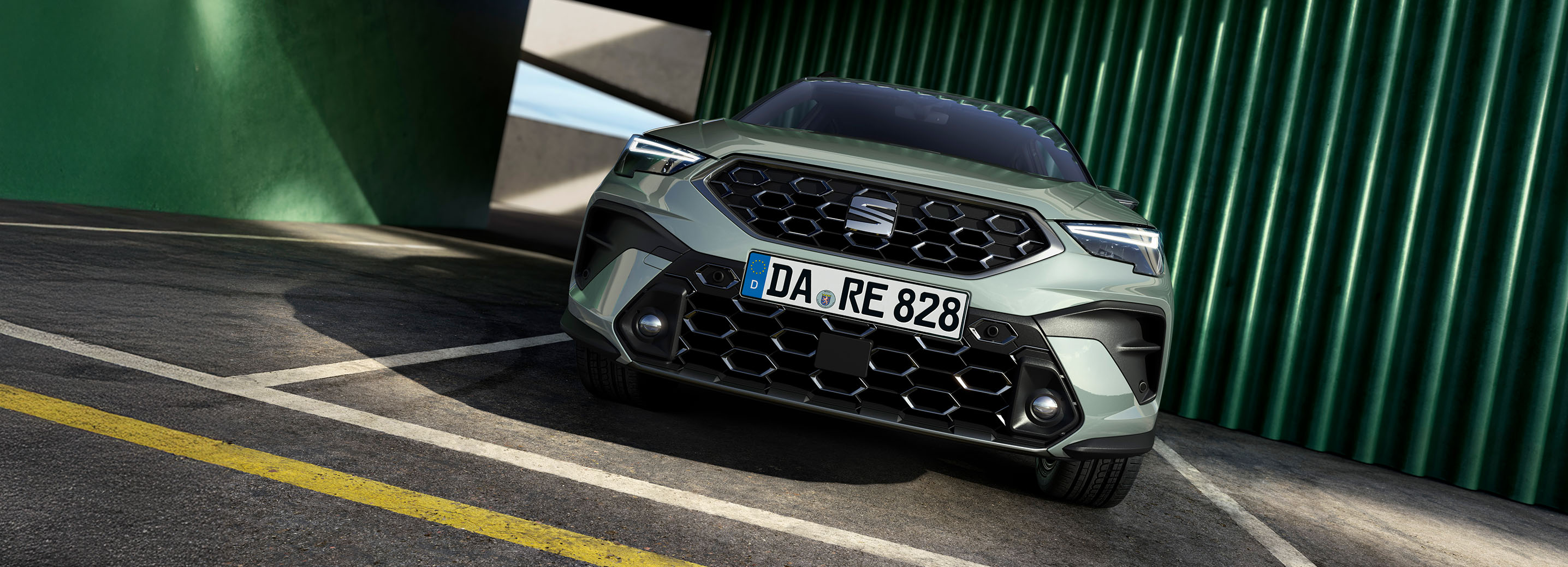 Frontalansicht eines SEAT Arona von vorne, geparkt auf einem Parkplatz vor einem modernen Gebäude mit gelben und grauen Elementen. 