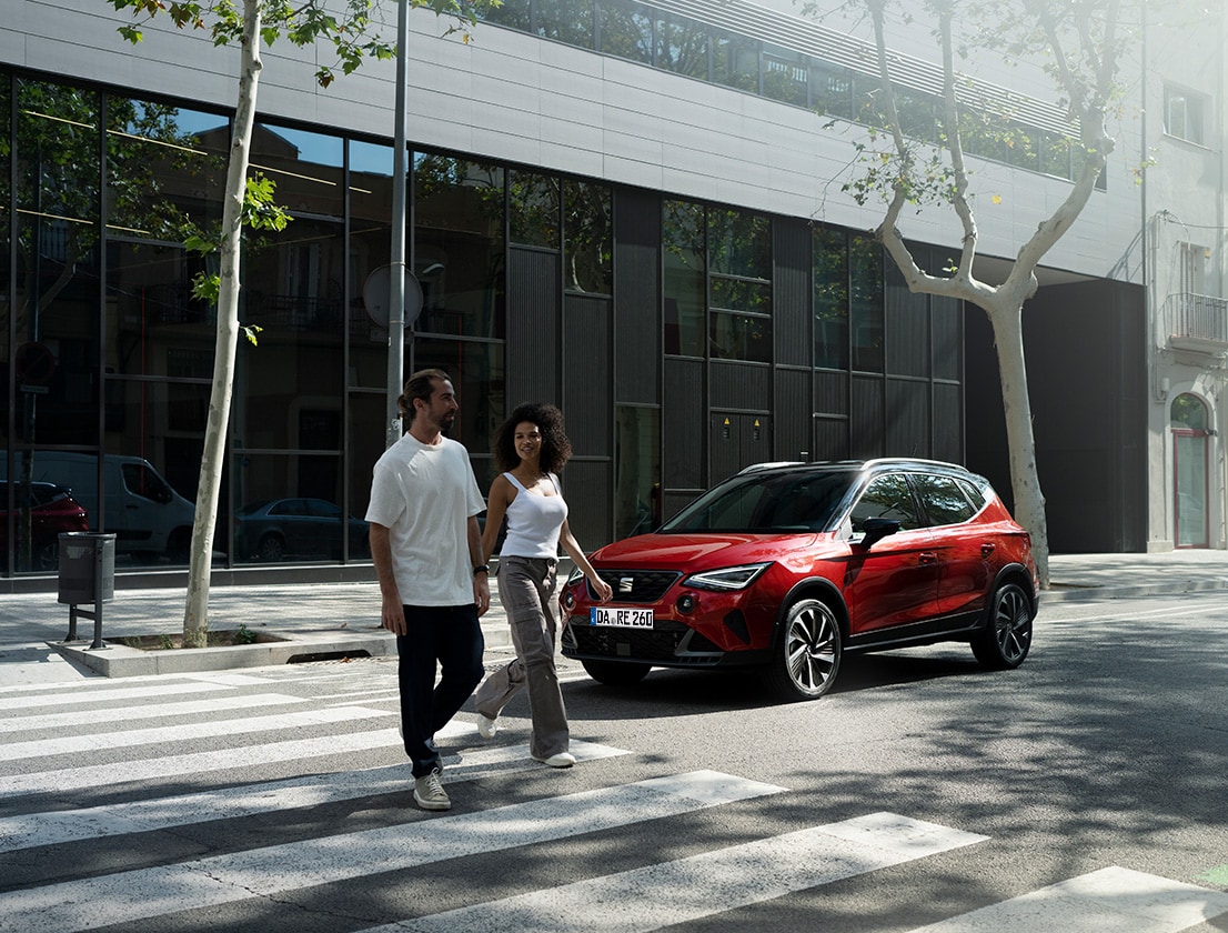 Ein roter SEAT Arona FR parkt am Straßenrand vor moderner Glasfassade, während ein junges Paar entspannt die Straße überquert.