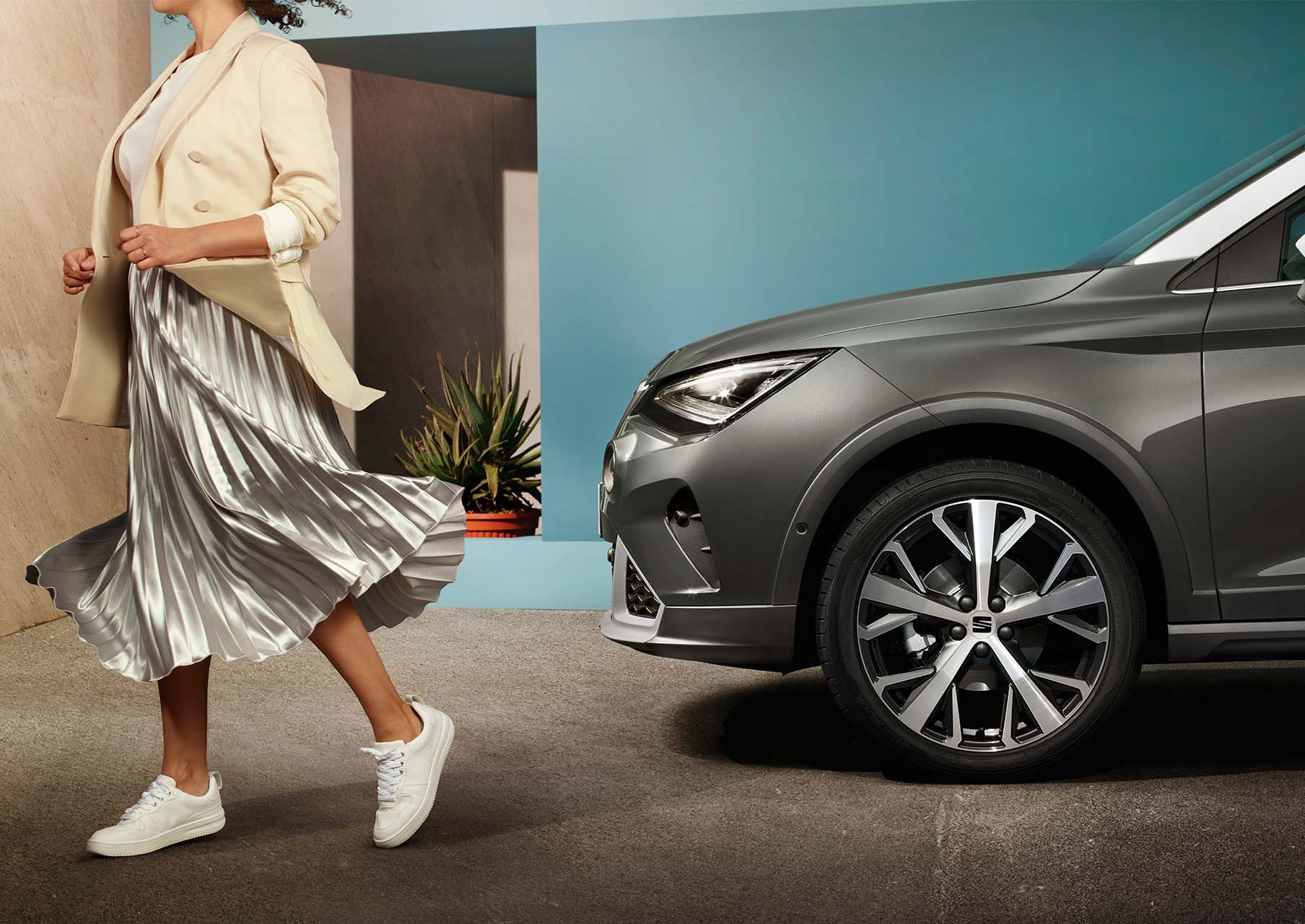 Eine Frau geht an der Front eines geparkten grauen SEAT Arona Xperience mit weißem Dach vorbei, im Hintergrund eine moderne Hauswand mit Pflanzen.