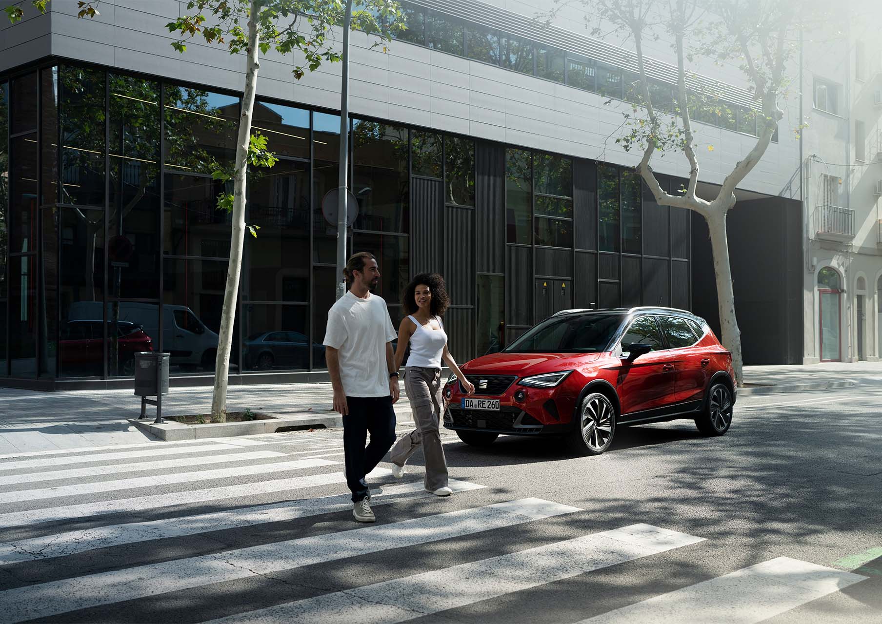 Eine Frau geht an der Front eines geparkten grauen SEAT Arona Xperience mit weißem Dach vorbei, im Hintergrund eine moderne Hauswand mit Pflanzen.