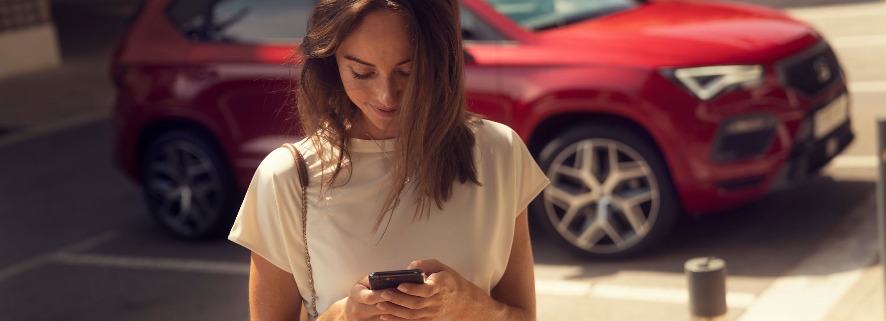 Frau blickt auf ihr Smartphone, im Hintergrund ein roter SEAT Ateca im Stadtumfeld