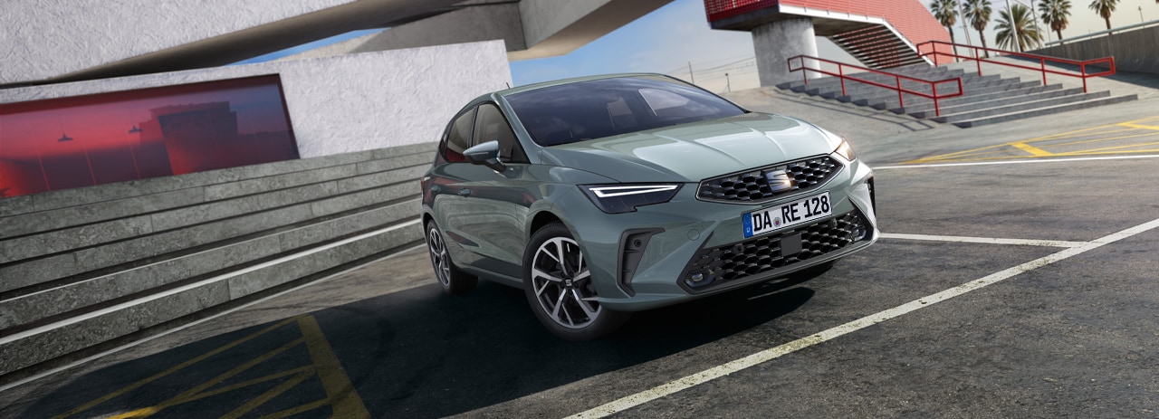 SEAT Ibiza in grün, frontale Außenansicht. Das Auto steht auf einem Parkplatz vor einer modernen Architektur mit Treppen und Palmen im Hintergrund. Das Kfz-Kennzeichen ist sichtbar