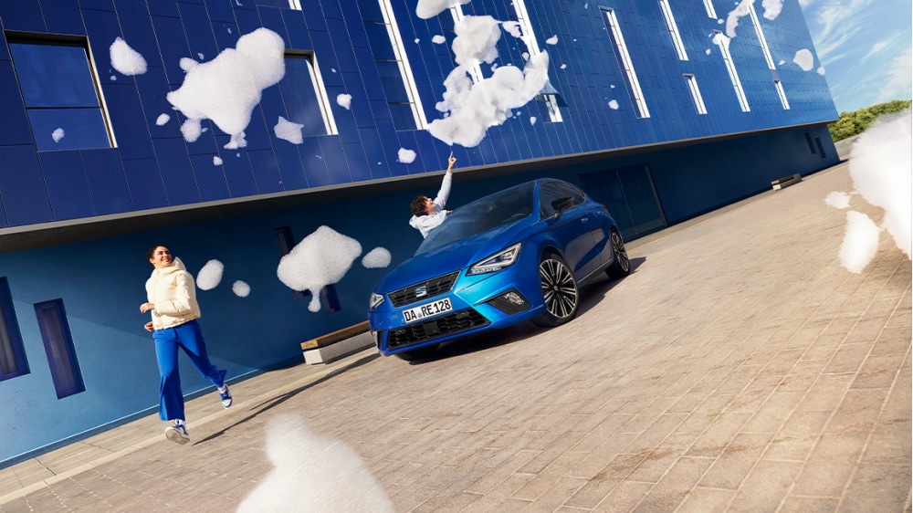 Ein blauer SEAT Ibiza steht vor einer blauen Wand. Von oben fallen Schaumwolken auf zwei Personen herab.