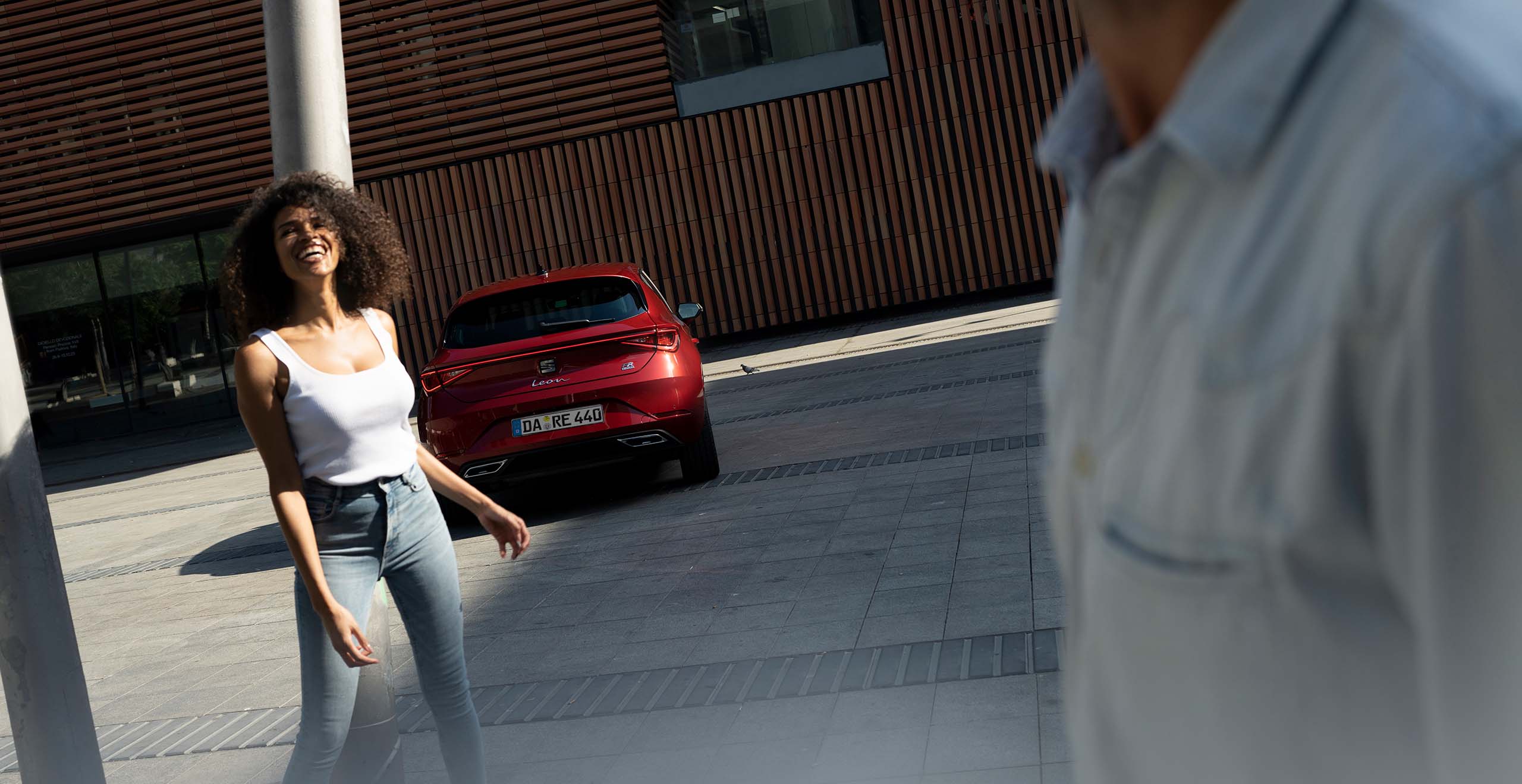 Junge Frau geht lächelnd über einen städtischen Platz, im Hintergrund steht ein roter SEAT Leon FR vor einem modernen Gebäude.
