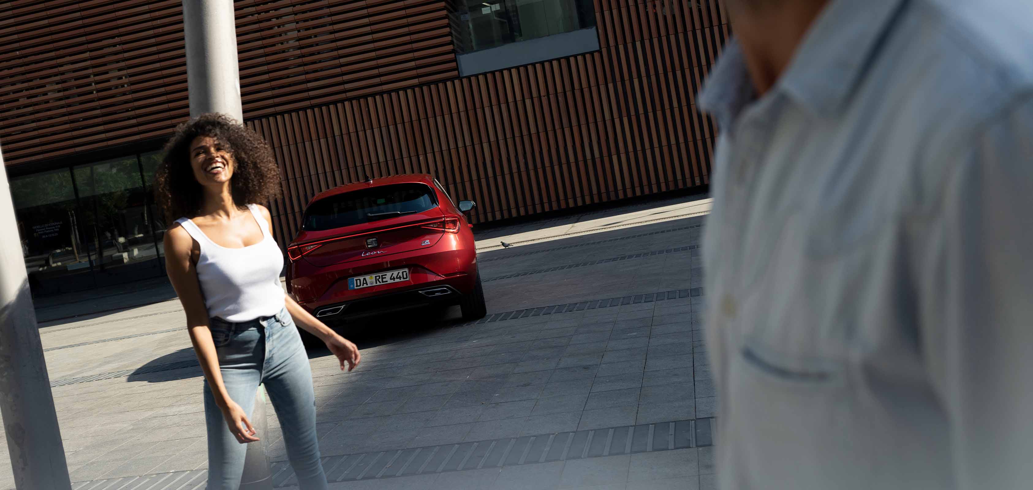 Junge Frau geht lächelnd über einen städtischen Platz, im Hintergrund steht ein roter SEAT Leon FR vor einem modernen Gebäude.