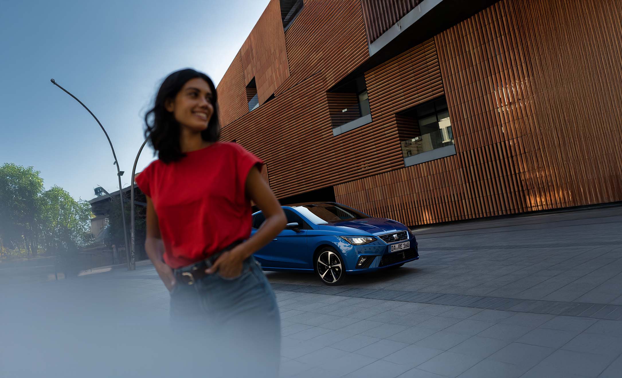 Junge Frau in rotem T-Shirt geht lächelnd vor einem geparkten blauen SEAT Ibiza FR entlang einer modernen Fassade mit Holzverkleidung.