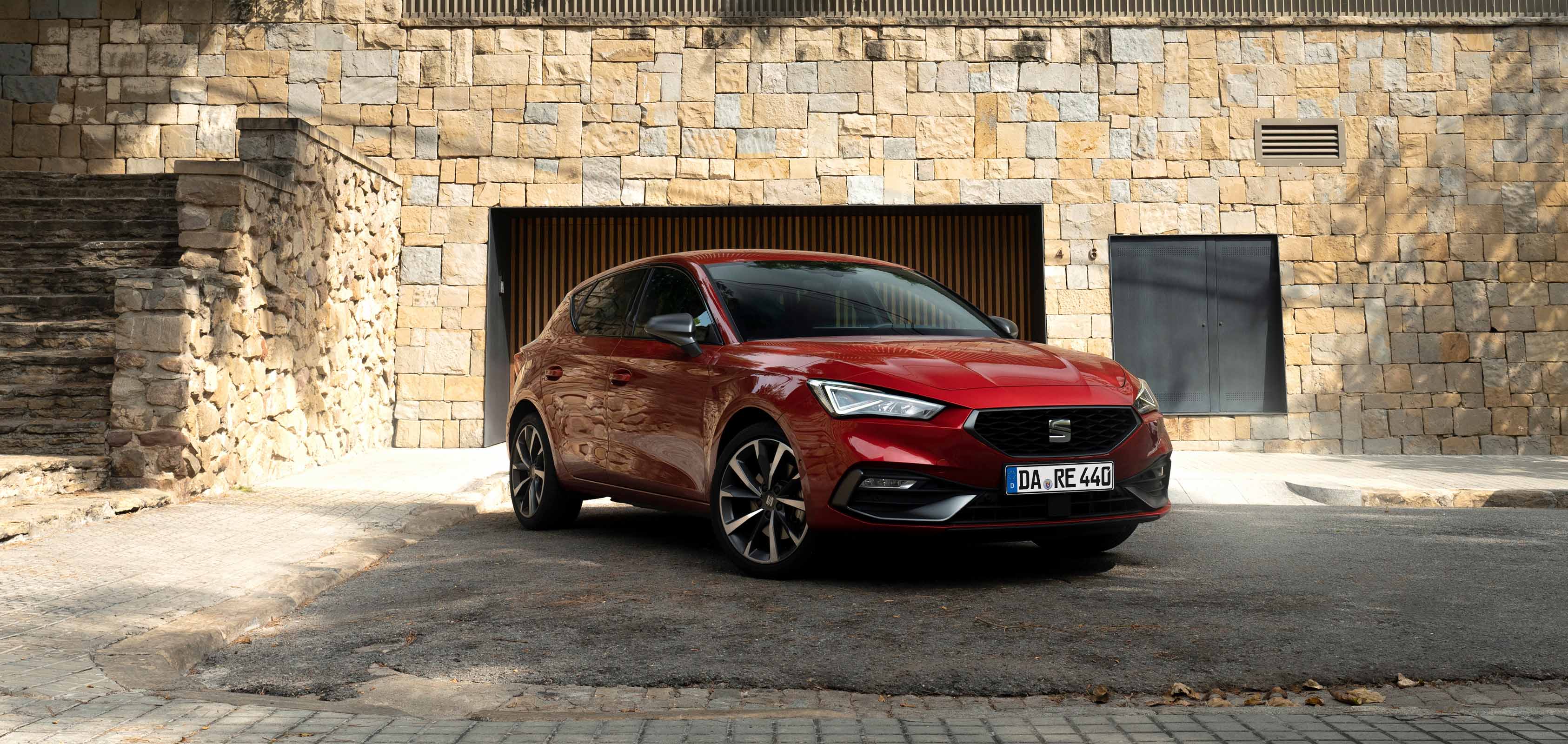 Ein roter SEAT Leon FR steht auf einer gepflasterten Einfahrt vor einem Wohnhaus mit Natursteinfassade und moderner Architektur.