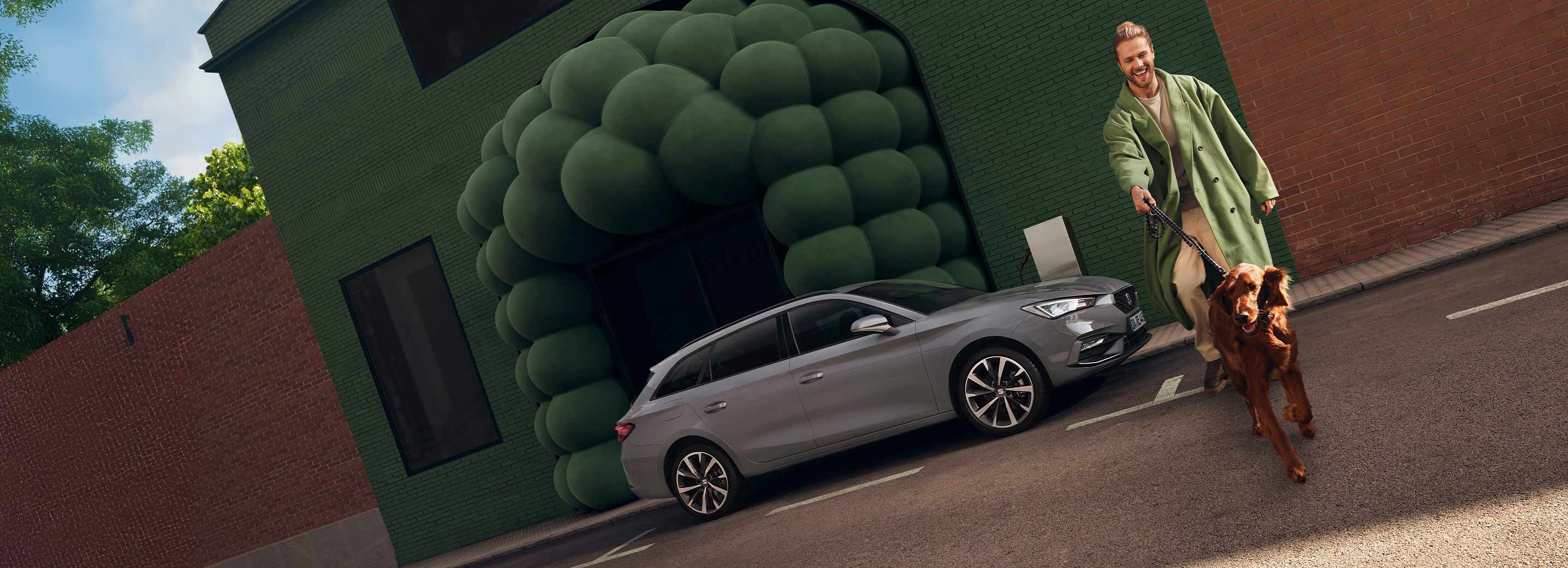 Ein Mann mit grünem Mantel und einem braunen Hund an der Leine läuft neben einem SEAT Leon Sportstourer über die Straße.