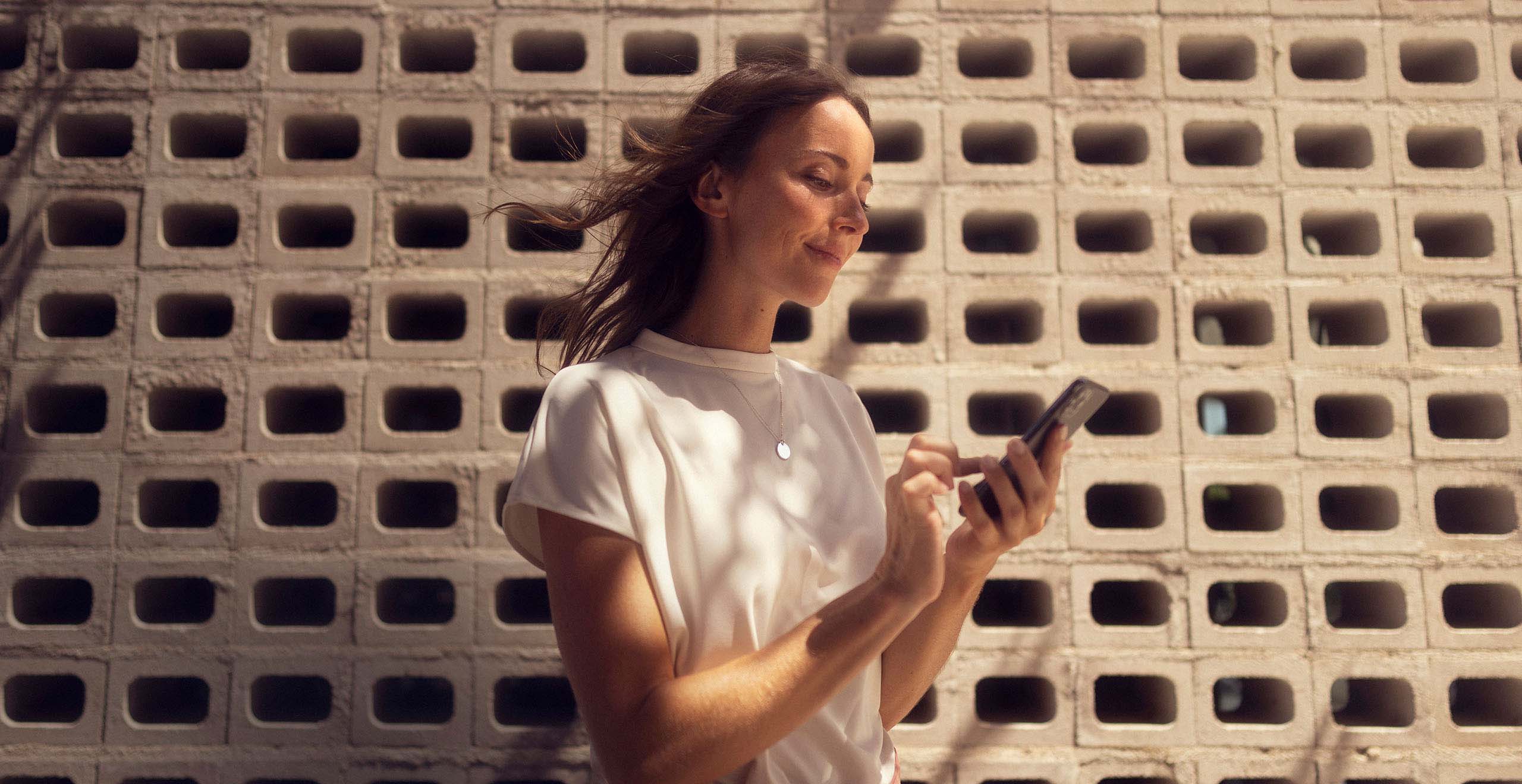Frau mit weißem Oberteil steht vor einer Betonwand und benutzt ein Smartphone.