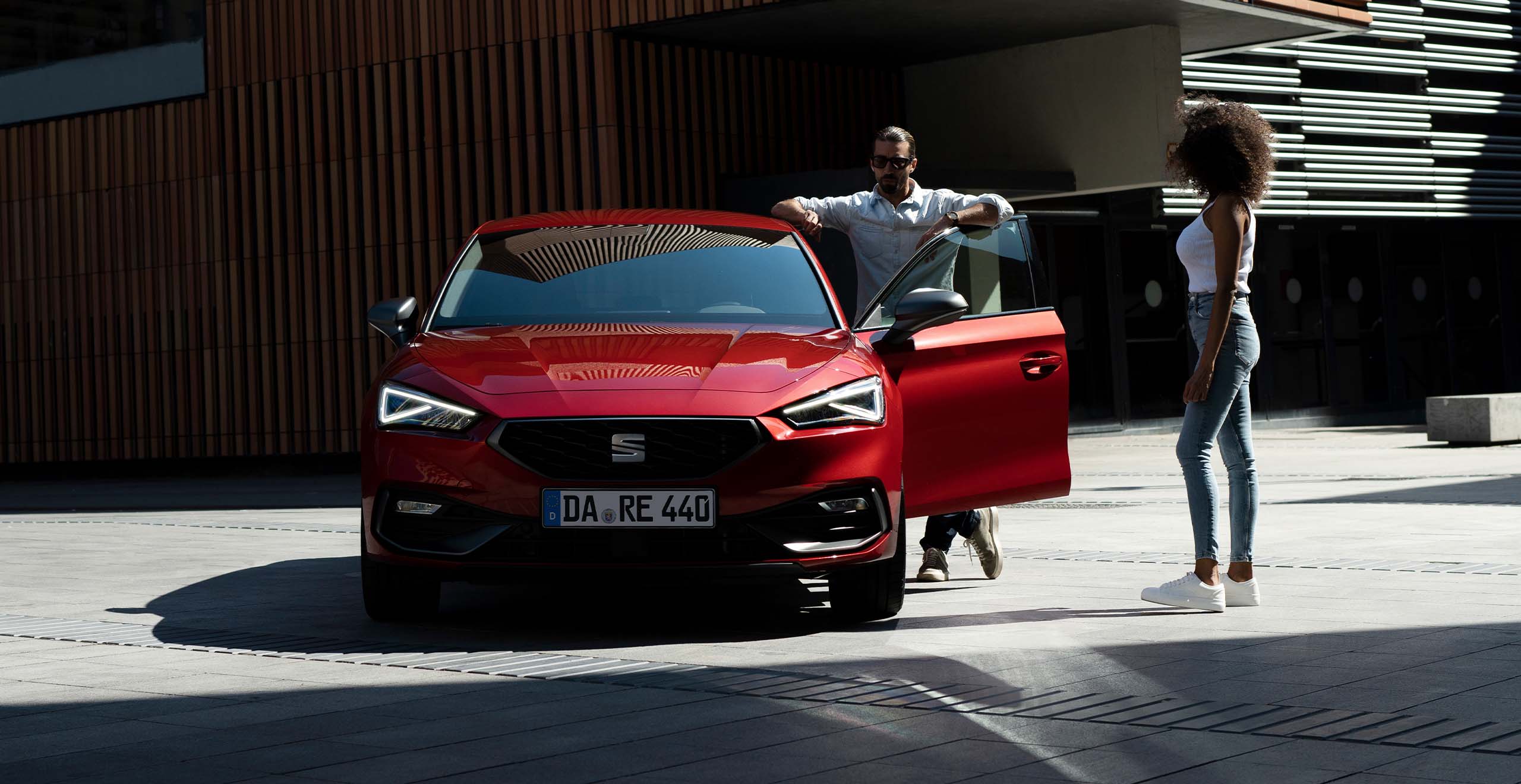 Roter SEAT Leon FR von vorne, geparkt auf einem modernen Platz; ein Mann steht an der geöffneten Fahrertür, eine Frau geht auf das Auto zu.