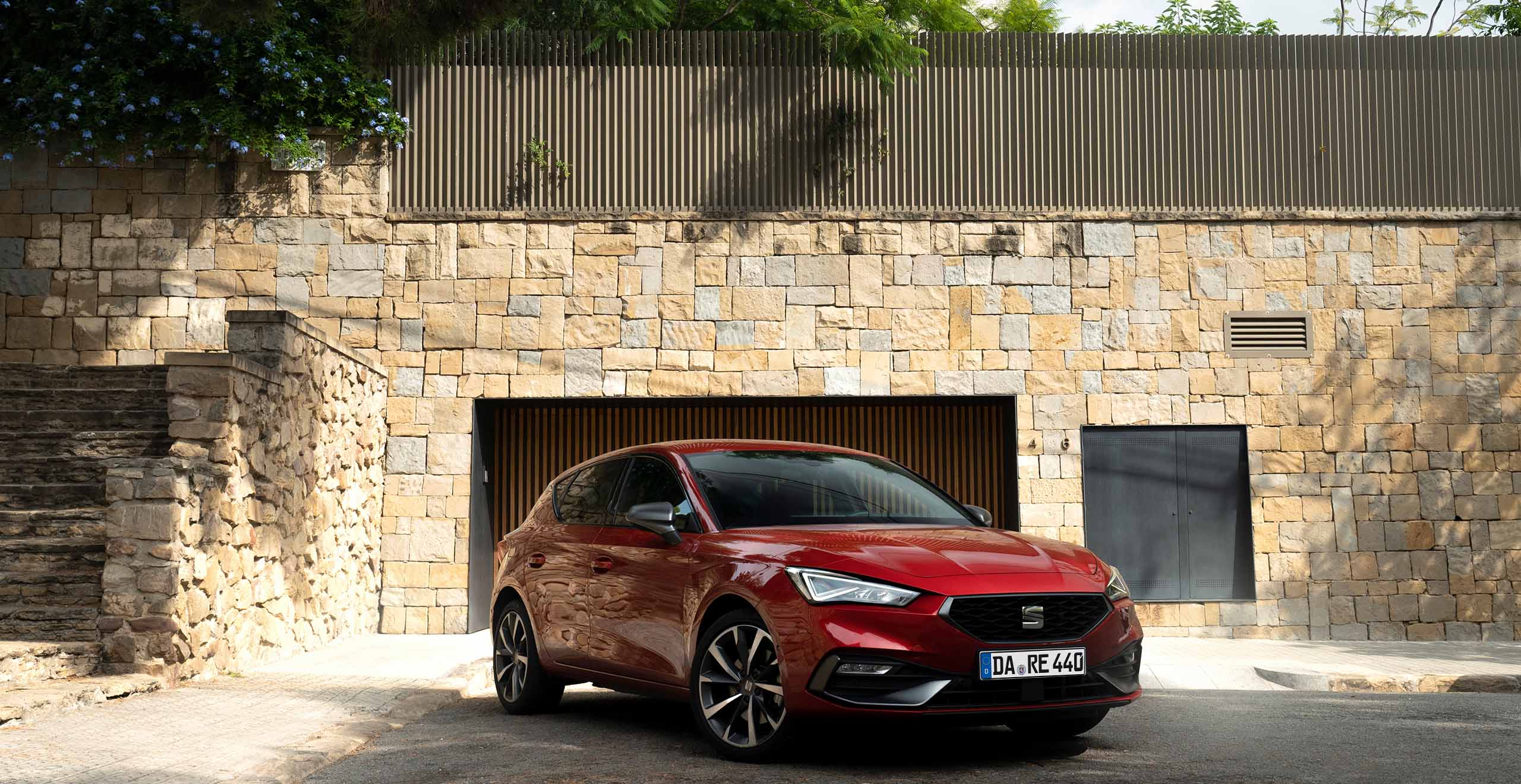 Ein roter SEAT Leon FR steht auf einer gepflasterten Einfahrt vor einem Wohnhaus mit Natursteinfassade und moderner Architektur.