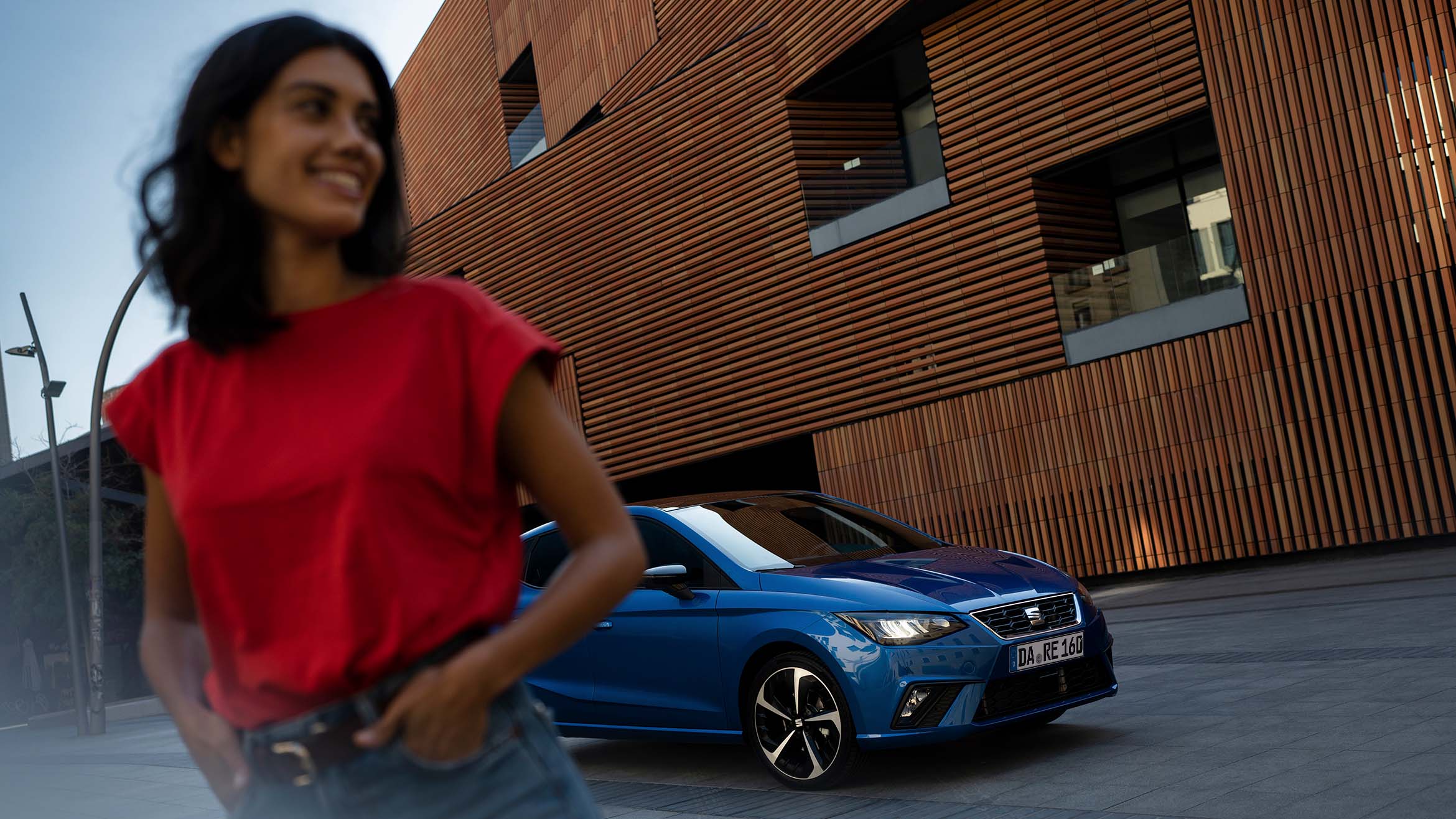 Junge Frau in rotem T-Shirt geht lächelnd vor einem geparkten blauen SEAT Ibiza FR entlang einer modernen Fassade mit Holzverkleidung.
