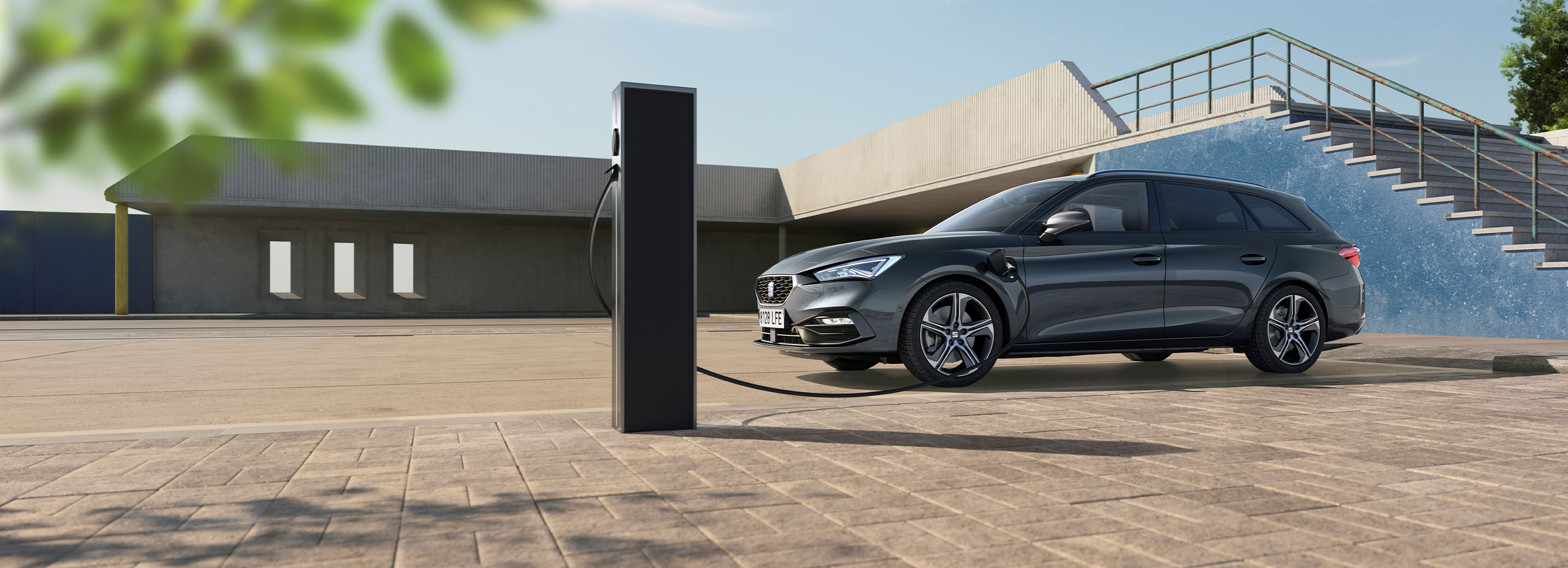 Ein SEAT Leon Sportstourer parkt auf einem sonnendurchfluteten Pflasterplatz und ist über ein Ladekabel mit einer Ladesäule verbunden. Im Hintergrund befinden sich moderne Betonarchitektur und eine Außentreppe vor einer leuchtend blauen Wand unter klarem Himmel.